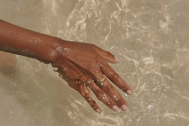 A woman's hand with dark brown skin is submerged in clear, shallow water.